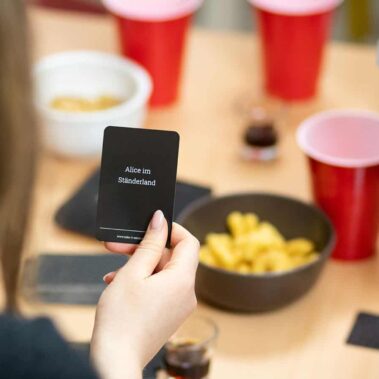 Take It Serious Porno-Editon: Spielkarte in der Hand einer Spielerin. Im Hintergrund sind Snacks und Partygetränke zu sehen.